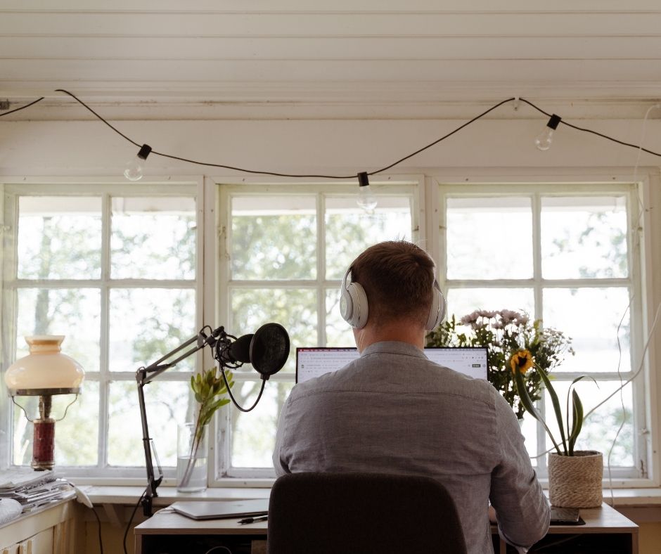 Mies työpöydän ääressä vaalean huvilan lasisella verannalla. Päässä kuulokkeet ja pöytään kiinnitetty studiomikrofoni.