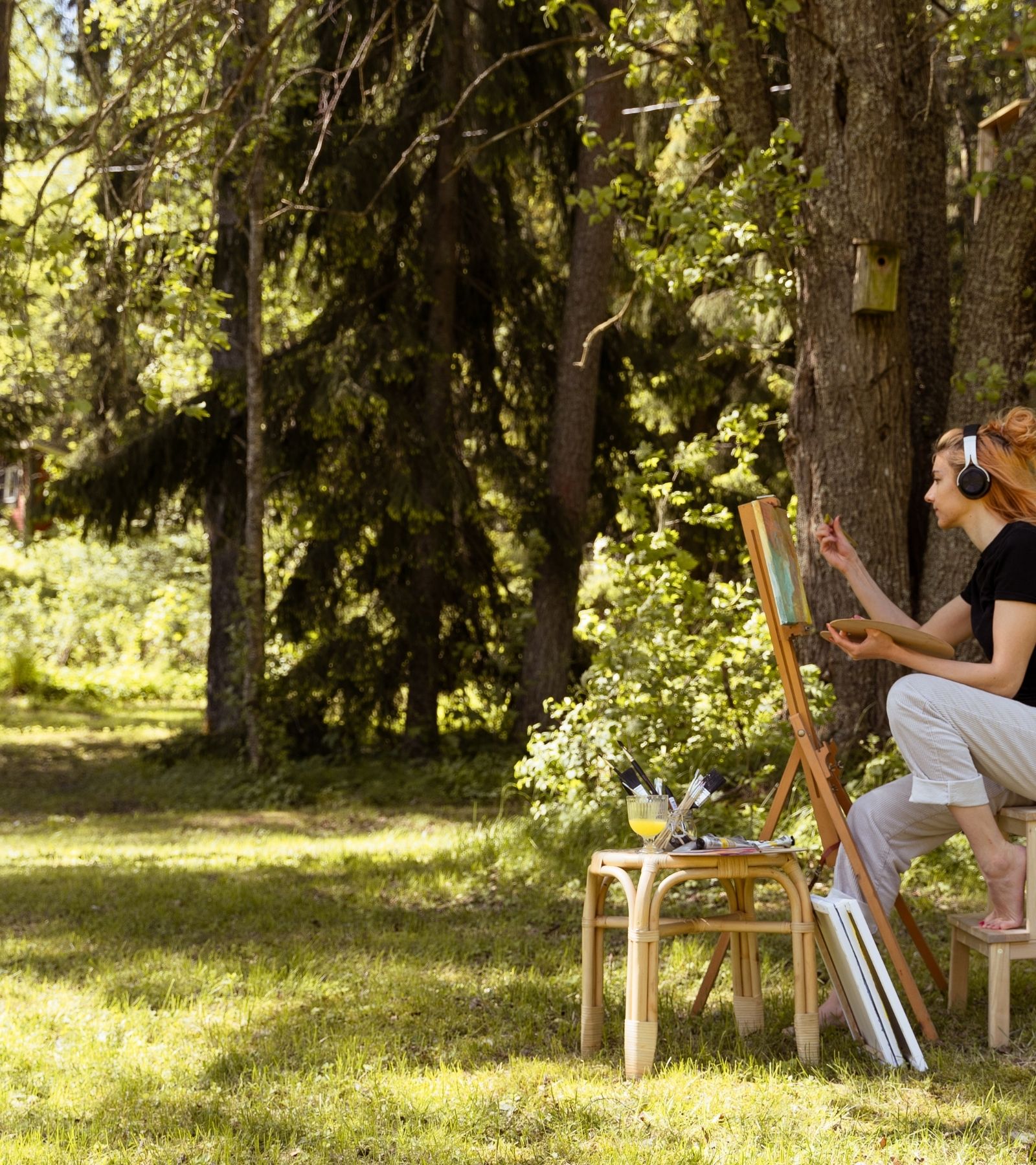 Nainen maalaa maalaustelineeseen kiinnitettyä canvastaulua ulkona kesällä. Päässä on kuulokkeet.