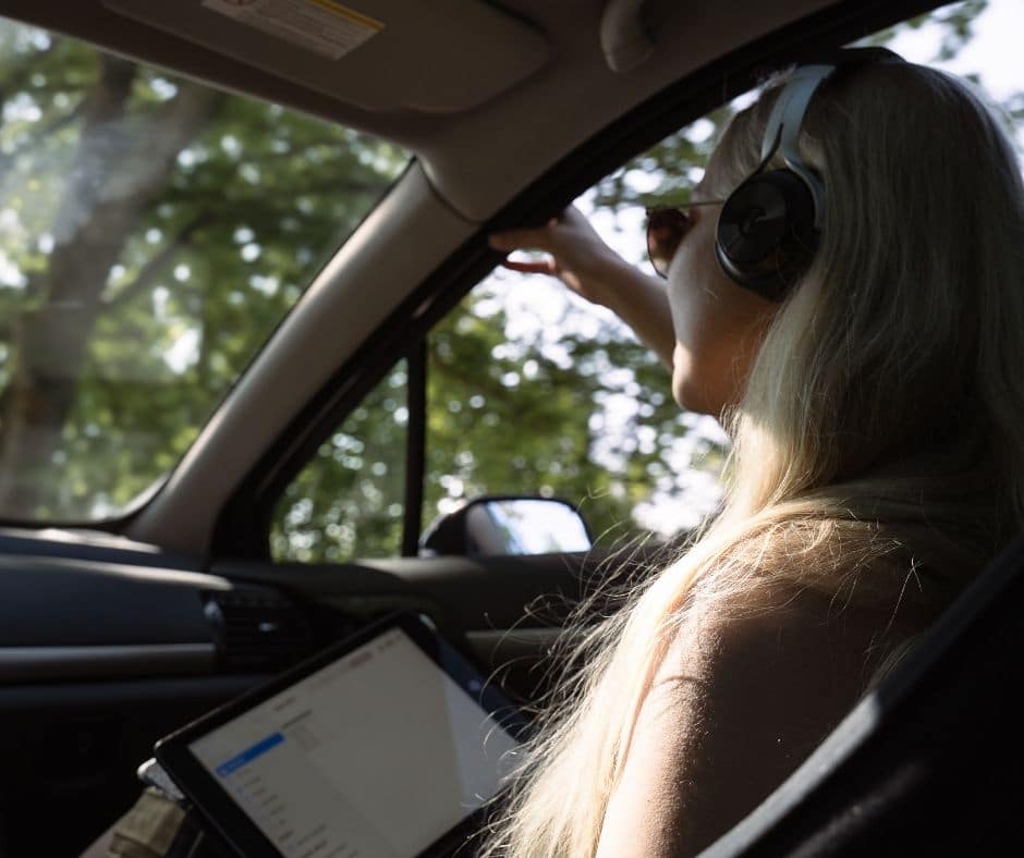 Nainen istuu auton etupenkillä kuulokkeet päässä ja tabletti sylissä.