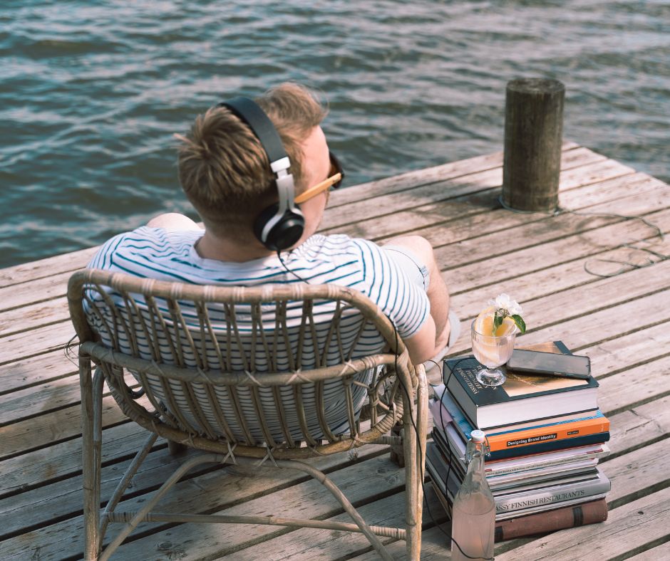 Mies istuu korituolissa laiturin nokassa ja katsoo merelle. Vieressä on kirjapino, jonka päällä älypuhelin. Miehellä on t-paita päällä ja kuulokkeet päässä.