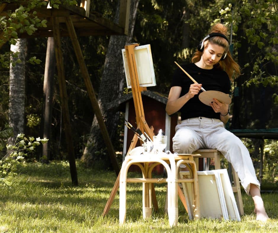 Nainen istuu kesällä puutarhassa taidemaalaustarvikkeiden kanssa. Päässä on kuulokkeet.
