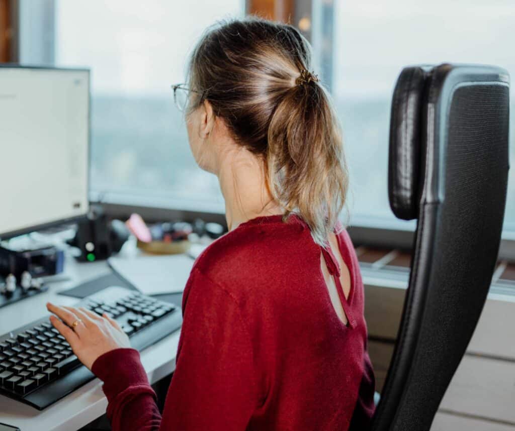 Nainen istuu toimistotuolissa tietokoneen edessä ja katsoo näyttöä. Toinen käsi on näppäimistöllä. Taustalla on isot ikkunat.
